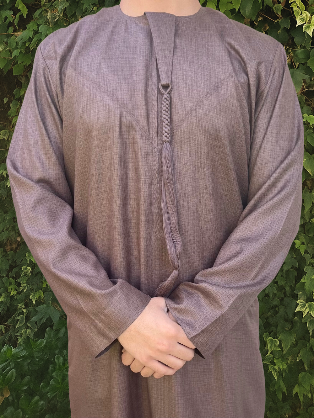 Person wearing a brown traditional outfit/thobe with a green leafy background