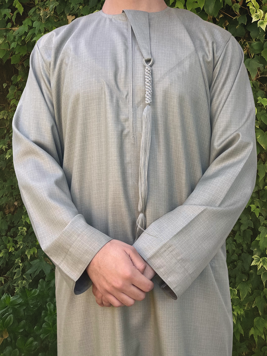 Person wearing a light green traditional outfit/thobe with a patterned tassel against a green leafy background
