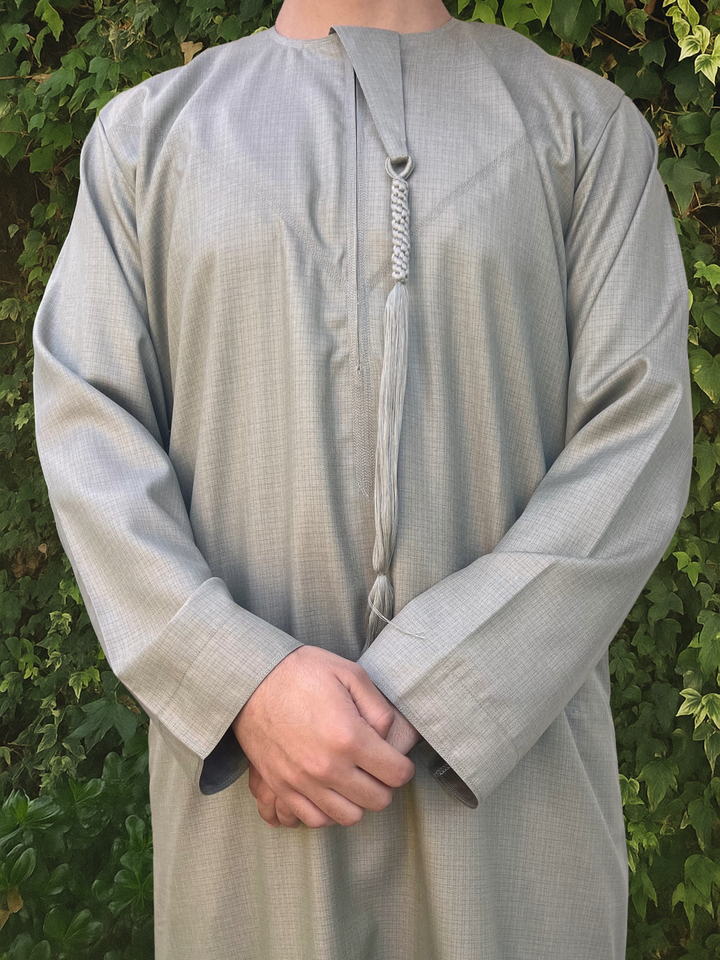 Person wearing a light green traditional outfit/thobe with a patterned tassel against a green leafy background