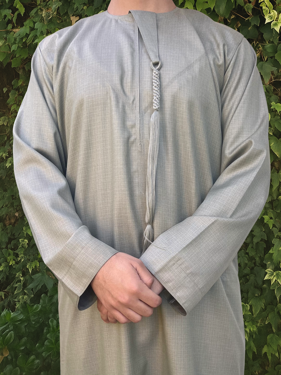 Person wearing a light green traditional outfit/thobe with a patterned tassel against a green leafy background