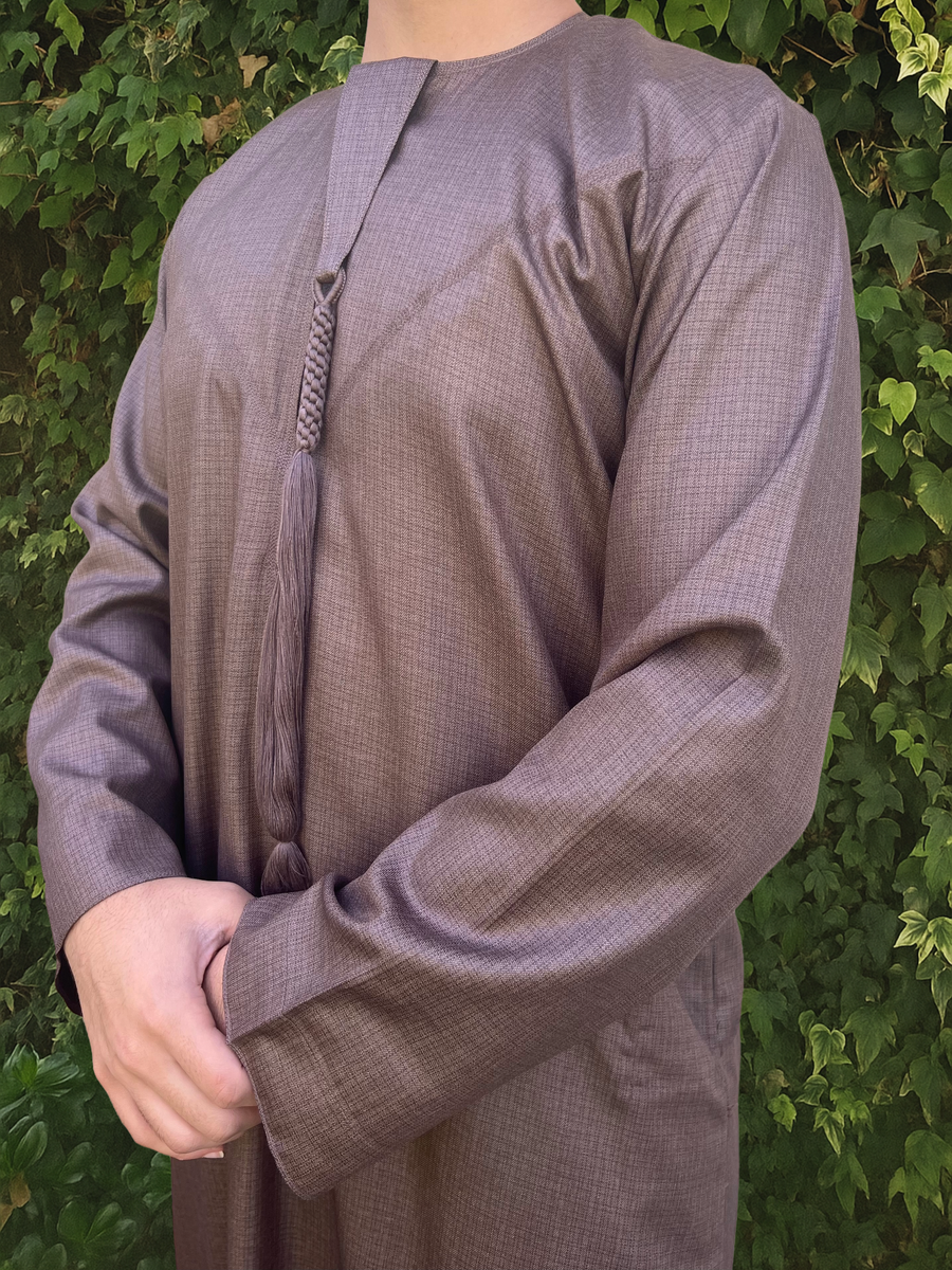 Person wearing a long-sleeve brown Emirati thobe with a tassel against a green leafy background